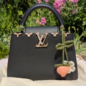 Op de afbeelding: Een zwarte leren handtas met gouden hardware en een gehaakte perzik charme. De tas heeft een bovenste handvat en een LV-logo aan de voorkant.