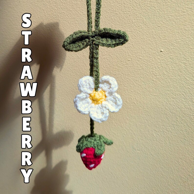 Op de afbeelding: Een gehaakte aardbei- en bloemenkrans met groene bladeren. De krans hangt aan een muur en de tekst "STRAWBERRY" is zichtbaar aan de linkerkant van de afbeelding.