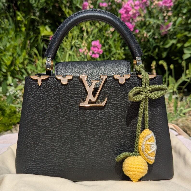 Op de afbeelding: Een zwarte leren handtas met gouden hardware en een gehaakte citroenbedel. De tas heeft een bovenste handvat en een afneembare schouderriem.