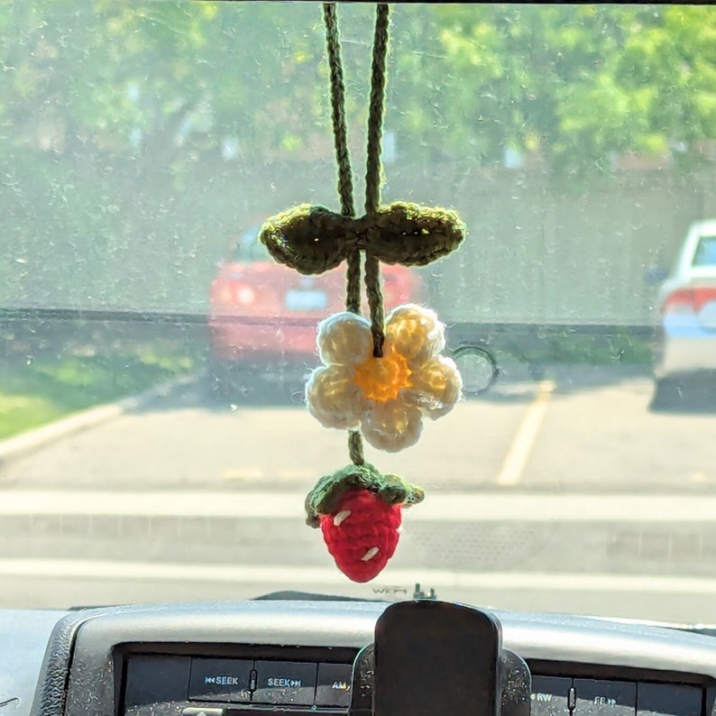 Op de afbeelding: Een gehaakte autohanger in de vorm van een aardbei, bloem en blad, hangend aan de achteruitkijkspiegel van een auto.