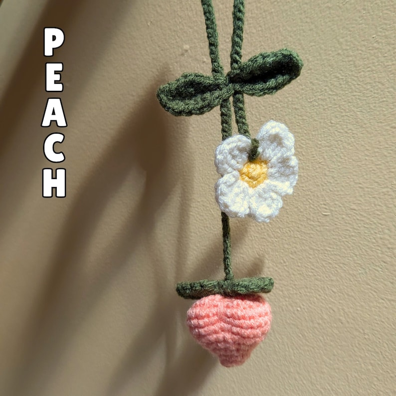 Op de afbeelding: Een gehaakte perzikbedel met een witte bloem en groene bladeren. De perzik is roze en de bloem heeft een gele kern.