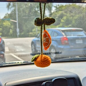 Op de afbeelding: Een gehaakte oranje hangende ornament met groene bladeren. De ornament is gevormd als een mandarijn met een schijfje sinaasappel aan de zijkant. De ornament hangt aan een achteruitkijkspiegel in een auto.