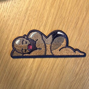 May include: Embroidered patch featuring a cartoon figure of a person lying down with their arms and legs outstretched. The figure is brown with a black outline and is wearing a black swimsuit. The patch is outlined in black.