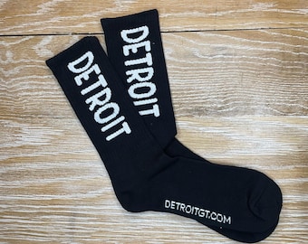 May include: A pair of black socks with white text that reads "DETROIT" on each sock. The socks have a white text label on the bottom that reads "DETROITGT.COM".