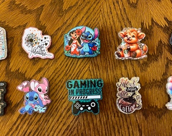 May include: Ten colorful, glittery, round pins with various designs. The pins feature images of coffee, a dandelion, Stitch and Angel from Lilo & Stitch, a cow, a video game controller, a coffee cup, and Stitch with balloons. The pins also include text such as "Coffee", "Just Breathe", "Gaming in Progress", "Life Begins After Coffee", and "Good Vibes Only".
