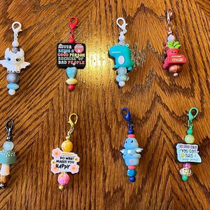 May include: A collection of 12 colorful zipper pulls with various charms and sayings. The charms include a dragon, a mushroom, a flower, a dinosaur, a pea pod, a minion, a plant, a cross, and a sign that says "You Got This".