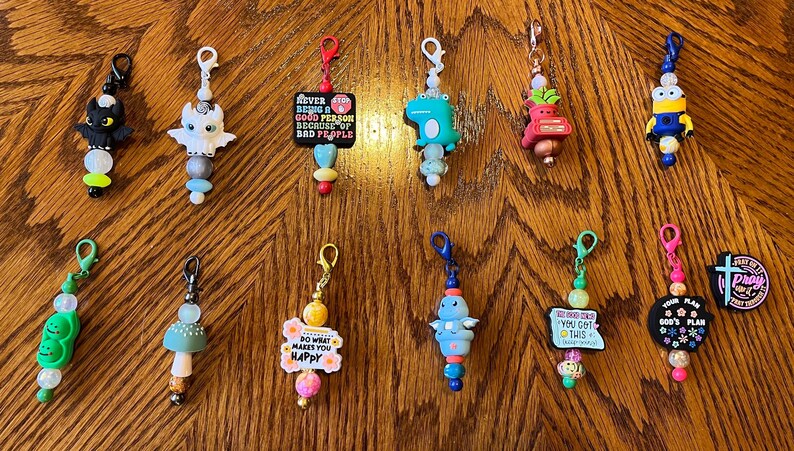 May include: A collection of 12 colorful zipper pulls with various charms and sayings. The charms include a dragon, a mushroom, a flower, a dinosaur, a pea pod, a minion, a plant, a cross, and a sign that says "You Got This".