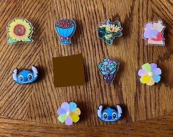 May include: Assortment of colorful, cartoon-themed, acrylic pins. The pins feature a sunflower, a bouquet of red roses, a bouquet of yellow flowers, a bouquet of blue flowers, a cartoon Stitch character, a cartoon Stitch character, a cartoon pig, a rainbow flower, and a yellow flower.