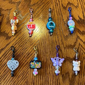 May include: A collection of 11 colorful zipper pulls with various charms, including a cartoon cat, a cartoon dog, a cross, a butterfly, a heart, a flower, and a red and white sign that says "Be Kind To Your Mind".
