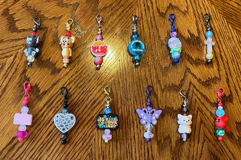 May include: A collection of 11 colorful zipper pulls with various charms, including a cartoon cat, a cartoon dog, a cross, a butterfly, a heart, a flower, and a red and white sign that says "Be Kind To Your Mind".
