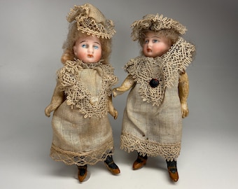 May include: Two antique bisque dolls. Each doll wears a tan dress with a lace collar and matching hat. The dolls have blonde hair, blue eyes, and painted facial features. They are standing side by side.