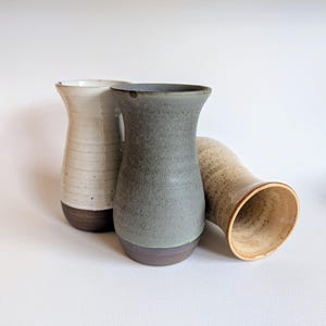 May include: Three ceramic vases with flared rims and tapered bodies. One vase is off-white with horizontal ridges, one is a matte sage green, and one is a speckled tan colour. Each vase features a dark brown, unglazed base section at the bottom.