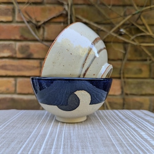 May include: Two ceramic bowls, one stacked on top of the other. The top bowl is white with brown stripes, and the bottom bowl is blue with white and black geometric designs.
