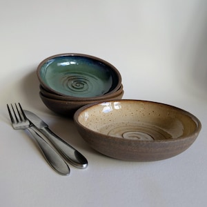 May include: A set of ceramic bowls with a dark brown exterior and a spiral pattern at the base. One bowl is beige with brown speckles, while the others feature a deep green and blue glaze. A stainless steel fork and knife are placed beside them.