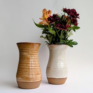 May include: Two ceramic vases, one with a textured brown finish and the other in speckled white, are displayed. The white vase holds a bouquet of orange, burgundy, and white flowers with green foliage. Both vases have a flared top and a brown base.