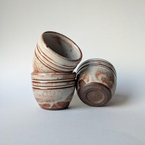 May include: Four small, handmade ceramic bowls. The bowls are a light beige colour with brown speckled patterns and horizontal brown stripes. One bowl is upside down, revealing a dark brown base. The bowls are stacked on a white surface.