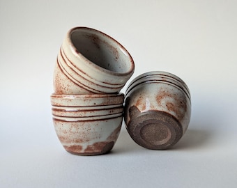 May include: Four small, handmade ceramic bowls. The bowls are a light beige colour with brown speckled patterns and horizontal brown stripes. One bowl is upside down, revealing a dark brown base. The bowls are stacked on a white surface.