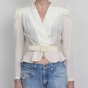 May include: Cream-colored wrap blouse with long sleeves and a matching belt. The blouse features a V-neck and a peplum-style hem. The model is wearing light blue denim jeans.