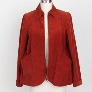 May include: A rust-coloured corduroy jacket featuring a pointed collar, long sleeves with folded cuffs, and four matching buttons down the front. The jacket includes two large, open patch pockets on the lower front panels.