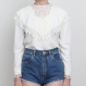Puede incluir: Una blusa blanca de manga larga con un gran cuello y puños de encaje ornamentados. La blusa se combina con unos shorts vaqueros azules. Los detalles de encaje añaden un toque vintage y femenino a la prenda.