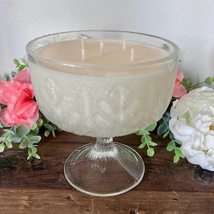 May include: A multi-wick candle in a clear glass pedestal bowl with a textured floral pattern on the exterior. The candle wax is a solid cream color, and the glass vessel features a wide rim and a sturdy circular base, showcasing a clear design.