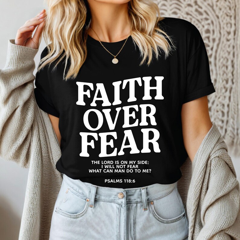 Może przedstawiać: Czarny T-shirt z białym napisem "FAITH OVER FEAR" i cytatem z Psalmu 118:6. Cytat brzmi: "Pan jest po mojej stronie: Nie będę się bał, co może mi zrobić człowiek?"