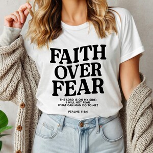 Może przedstawiać: Biała koszulka z czarnym nadrukiem "FAITH OVER FEAR" i wersetem "THE LORD IS ON MY SIDE: I WILL NOT FEAR WHAT CAN MAN DO TO ME? PSALMS 118:6"
