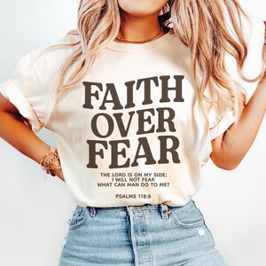 Może przedstawiać: Biały T-shirt z napisem "FAITH OVER FEAR" w brązowych literach. Na koszulce znajduje się r&oacute;wnież tekst "THE LORD IS ON MY SIDE: I WILL NOT FEAR WHAT CAN MAN DO TO ME? PSALMS 118:6" w mniejszych brązowych literach.