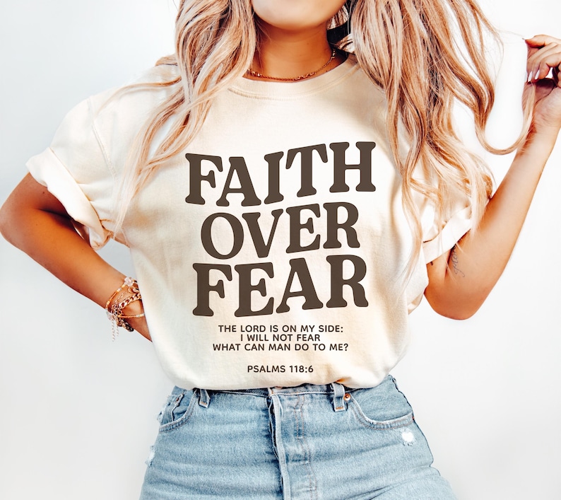 Może przedstawiać: Biały T-shirt z napisem "FAITH OVER FEAR" w brązowych literach. Na koszulce znajduje się r&oacute;wnież tekst "THE LORD IS ON MY SIDE: I WILL NOT FEAR WHAT CAN MAN DO TO ME? PSALMS 118:6" w mniejszych brązowych literach.