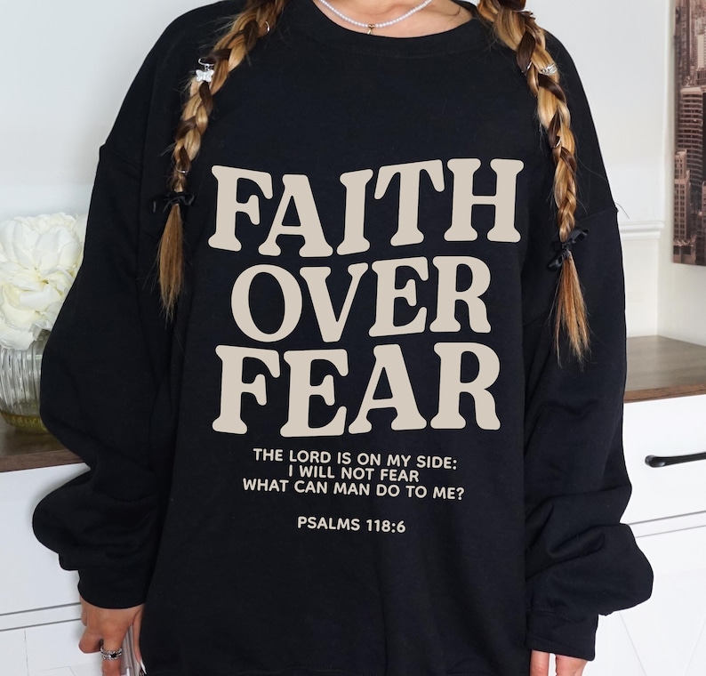 Może przedstawiać: Czarny sweter z napisem "FAITH OVER FEAR" w beżowych literach. Poniżej gł&oacute;wnego napisu znajduje się mniejszy napis w beżowych literach: "THE LORD IS ON MY SIDE: I WILL NOT FEAR WHAT CAN MAN DO TO ME? PSALMS 118:6".