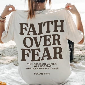 Może przedstawiać: Biała koszulka z napisem "FAITH OVER FEAR" w brązowych literach. Poniżej gł&oacute;wnego tekstu, mniejszymi brązowymi literami, nadrukowany jest tekst "THE LORD IS ON MY SIDE: I WILL NOT FEAR WHAT CAN MAN DO TO ME? PSALMS 118:6".