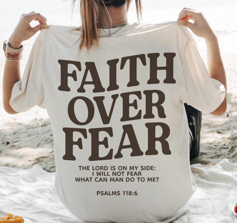 Może przedstawiać: Biała koszulka z napisem "FAITH OVER FEAR" w brązowych literach. Poniżej gł&oacute;wnego tekstu, mniejszymi brązowymi literami, nadrukowany jest tekst "THE LORD IS ON MY SIDE: I WILL NOT FEAR WHAT CAN MAN DO TO ME? PSALMS 118:6".