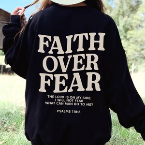 Może przedstawiać: Czarna bluza z nadrukiem "FAITH OVER FEAR" w kolorze białym na plecach. Pod gł&oacute;wnym tekstem znajduje się napis "THE LORD IS ON MY SIDE: I WILL NOT FEAR WHAT CAN MAN DO TO ME? PSALMS 118:6".