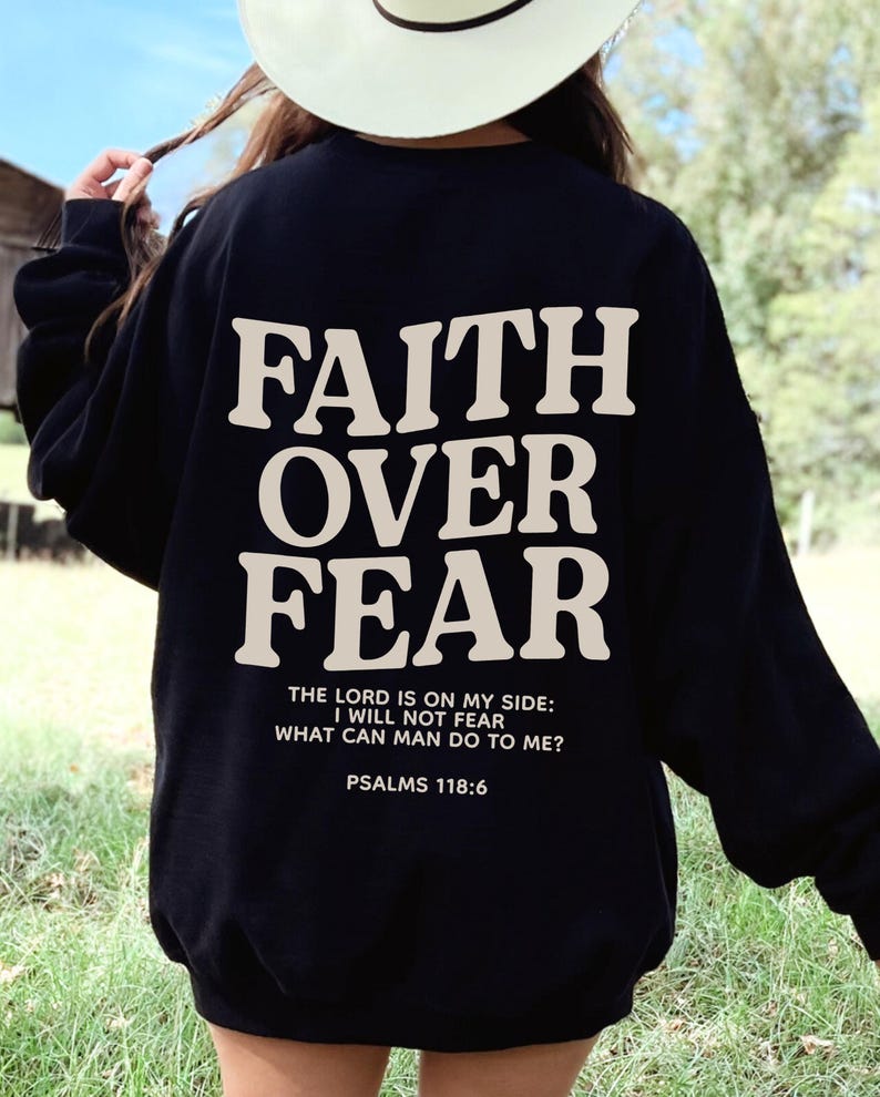 Może przedstawiać: Czarna bluza z nadrukiem "FAITH OVER FEAR" w kolorze białym na plecach. Pod gł&oacute;wnym tekstem znajduje się napis "THE LORD IS ON MY SIDE: I WILL NOT FEAR WHAT CAN MAN DO TO ME? PSALMS 118:6".
