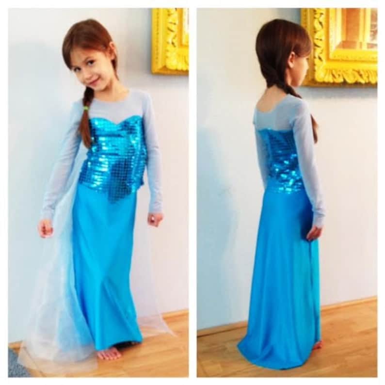 Puede incluir: Una ni&ntilde;a peque&ntilde;a con un vestido azul y plateado con lentejuelas, con una falda larga azul y una capa transparente de color azul claro. El vestido tiene un escote de coraz&oacute;n y mangas largas.