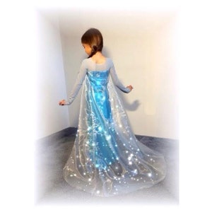 Puede incluir: Un vestido de princesa azul claro con una larga capa de tul fluida. La capa tiene un dise&ntilde;o brillante con purpurina blanca y detalles en azul claro. El vestido tiene mangas largas y un corpi&ntilde;o ajustado.