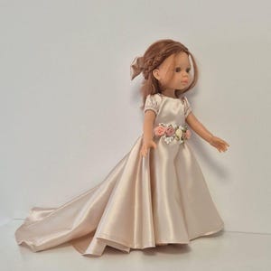Puede incluir: Muñeca con un vestido de satén color champán con una cola fluida. El vestido presenta un adorno floral en la cintura. La muñeca tiene cabello castaño rojizo peinado con un lazo.