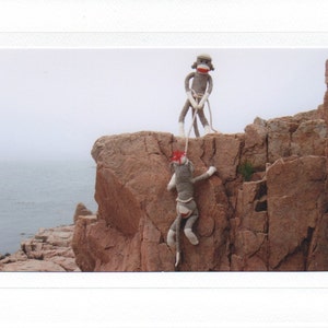 May include: Two gray sock monkeys are climbing a rocky cliff. The monkey on top is holding a rope and the monkey below is hanging from the rope. The background is a blue ocean and a rocky shore.