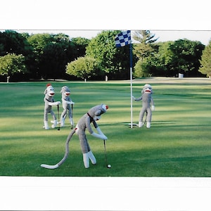 May include: Four gray sock monkey figures playing golf on a green golf course. One monkey is putting a golf ball, while the other three are holding golf clubs.