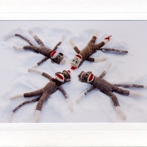 May include: Four brown sock monkeys making snow angels in the snow. The monkeys are wearing red and white hats.