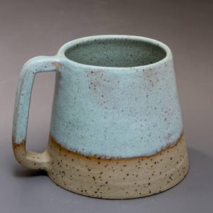 May include: A ceramic mug with a tapered silhouette and a flat-sided handle. The upper portion features a speckled light blue glaze, while the lower base is a speckled tan clay. The interior is finished in a matching speckled light blue glaze.