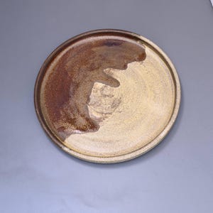 May include: A round ceramic plate featuring a speckled beige base with a prominent, dark brown glazed section that sweeps across one side. The plate has a slightly raised rim and a textured, earthy appearance with visible variations in colour.