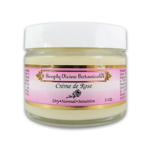 Puede incluir: Un tarro de cristal de Crème de Rose de Simply Divine Botanicals. El tarro tiene una tapa blanca y una etiqueta rosa con detalles dorados. La etiqueta indica que la crema es para pieles secas, normales y sensibles. El tarro contiene 57 gramos de crema.