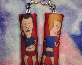 May include: A pair of plush keyrings featuring cartoon characters. Each keyring has a red rectangular base with a cartoon image. One character wears a blue shirt with "AC/DC" text, the other wears a blue shirt. Both have black shorts. The keyrings are connected by heart-shaped clasps.