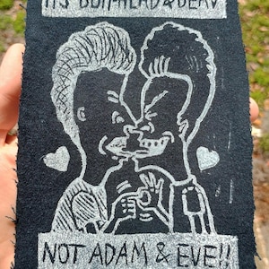 Könnte beinhalten: Schwarzer Aufnäher mit einer weißen Illustration von Beavis und Butt-Head beim Küssen, mit dem Text "IT'S BUTTHEAD & BEAV" darüber und "NOT ADAM & EVE!!" darunter. Zwei kleine Herzen sind ebenfalls im Design enthalten.