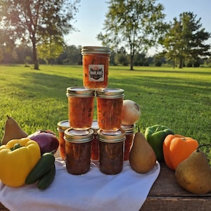 May include: A stack of six glass jars filled with a red and orange salsa. The jars are labeled "Bright Acres". The jars are surrounded by various vegetables, including yellow and orange bell peppers, green bell peppers, pears, onions, and green jalapenos.