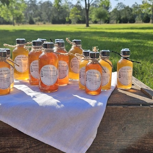 May include: A collection of small glass bottles filled with a light amber liquid, arranged on a wooden crate covered with a white cloth. The bottles have black lids and labels with white text that reads "Real North Vanilla Honey".