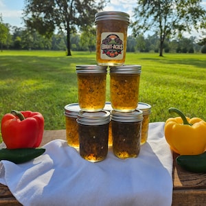 May include: Six glass jars of pepper jelly stacked in a pyramid shape. The jars are filled with a golden yellow jelly and have silver lids. The jars are labeled "Bright Acres".  A red bell pepper, a yellow bell pepper, and two green jalapeno peppers are on a wooden surface in front of the jars.
