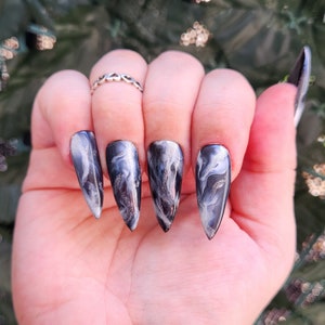 Op de afbeelding: Close-up van een hand met vijf lange, puntige nagels. Elke nagel is geverfd met een zwart en wit marmerpatroon.