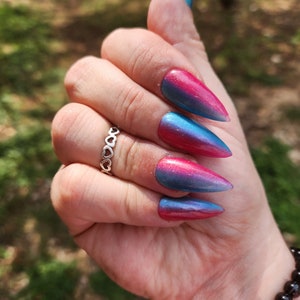 May include: Close-up of a hand with long, pointed, blue and pink ombre nails. The nails have a shimmery finish and are accented with a silver ring.